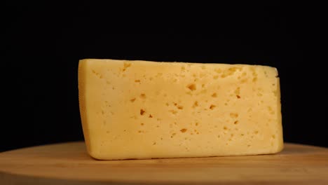 slow rotation of piece of yellow cheese with holes on wooden board on black background.