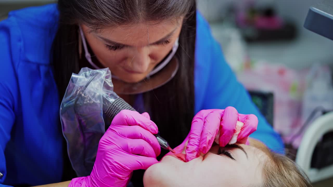 Lips tattoo. Cosmetologist in protective gloves applying permanent make-up on lips. Professional tattooist working with special tattooer machine.