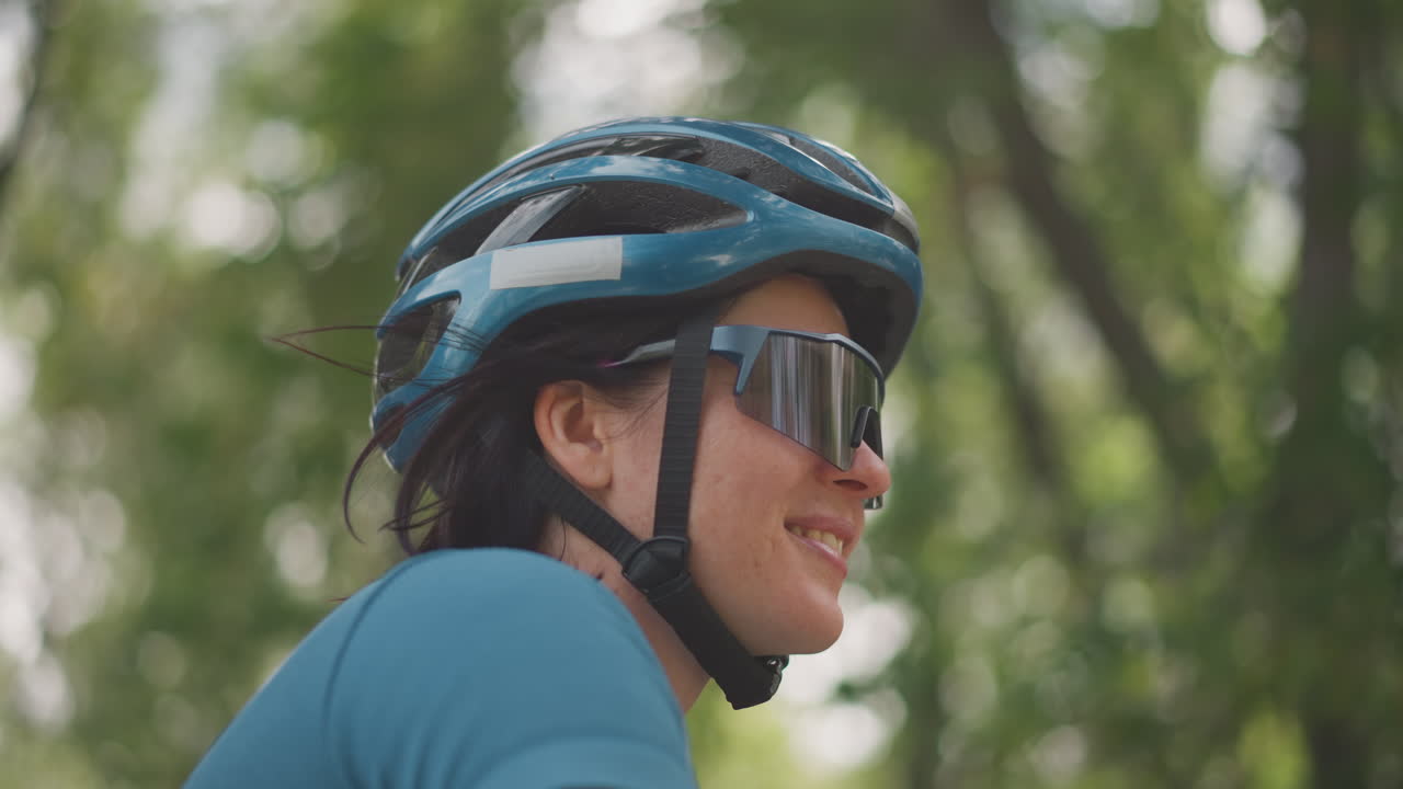 asian woman cyclist wearing helmet and sunglasses, smiling during solo forest ride, blue jersey, dappled sunlight through trees, calm expression, slow motion profile, closeup portrait ideal