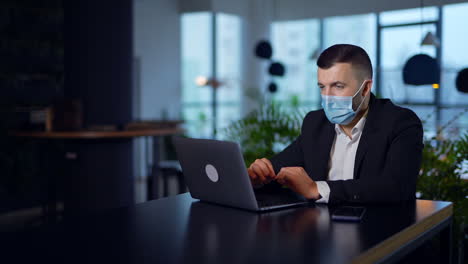 serious man wearing mask sitting at the desk. businessman printing something at his laptop. blurred backdrop.