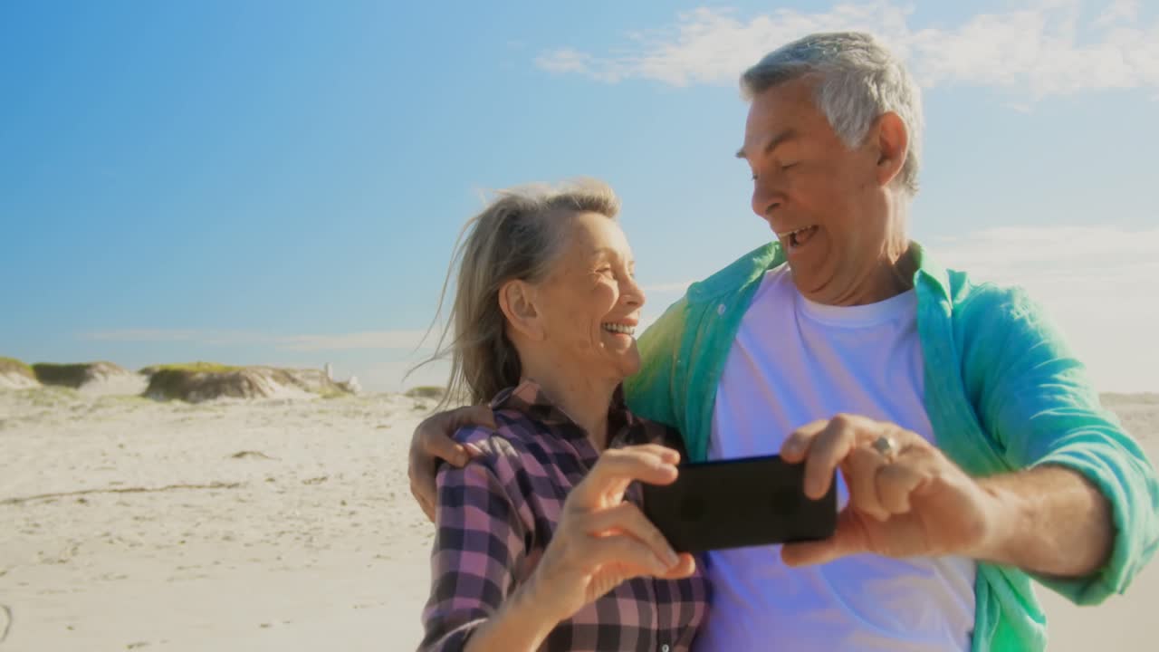 vista frontal de una pareja caucásica senior activa tomando una selfie con un teléfono móvil en la playa 4k