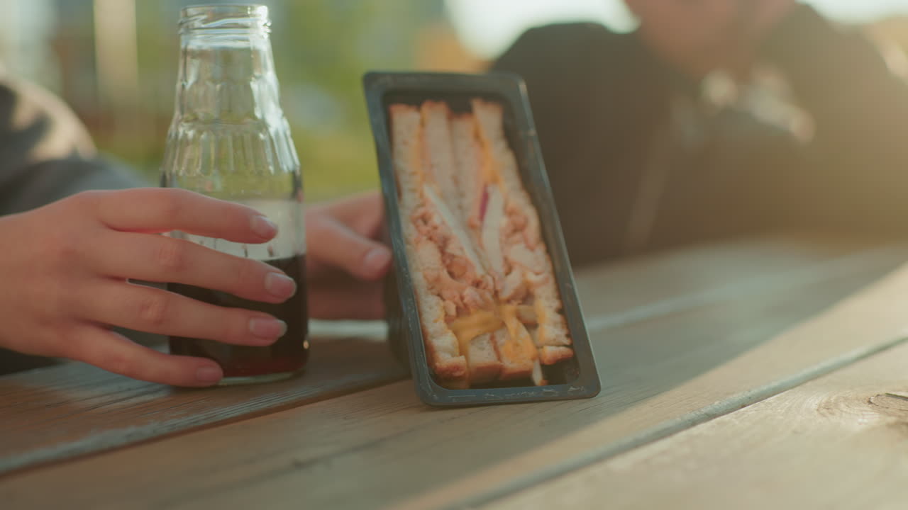 Close up of adult placing juice bottle and sandwich pack with sliced bread on wooden table for kid sitting nearby, with warm sunlight glow creating cozy outdoor atmosphere