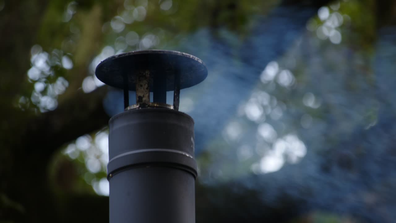 tiro cercano de una chimenea de chimenea de metal con humo ligero, árbol grande detrás