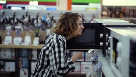 Side-view-of-a-young-curly-haired-woman-in-plaid-shirt-chooses-a-microwave-oven-in-a-consumer-electronics-supermarket.-Opens-the-door-and-looking-inside-the-oven