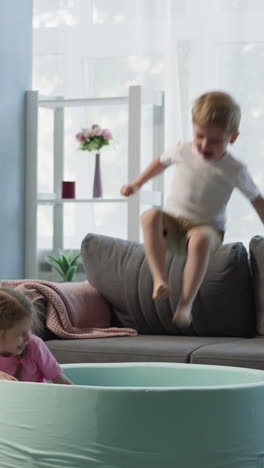 couple of grateful children jumps into dry pool from sofa. toddler boy and preschooler girl siblings play throwing colorful plastic balls with laugh