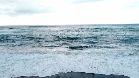 Pull-drone-shot-of-a-blonde-girl-walking-in-a-black-sand-beach-in-canary-island-with-rough-ocean-in-a-cloudy-moody-day