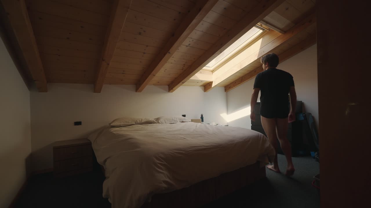 Uomo che entra in camera da letto, guarda la luce del sole dalla finestra Velux sul tetto e poi si siede sul letto