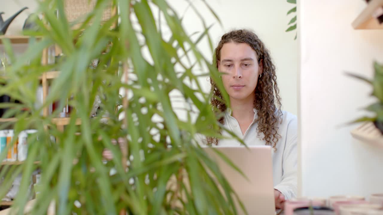 La telecamera si allontana dalla pianta rivelando una persona non binaria afroamericana che usa un laptop alla scrivania per lavoro.