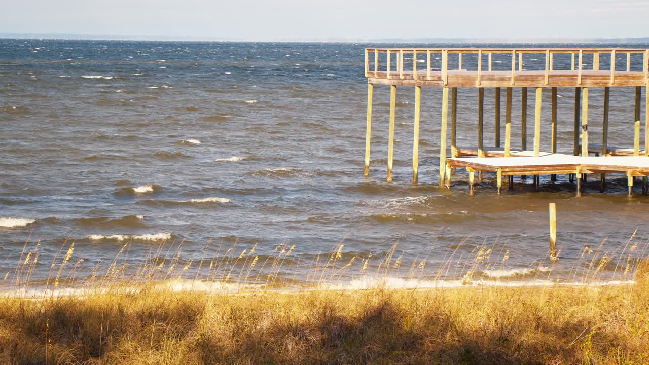 Rough ocean waves and a breezy day in Gulf Breeze, FL. A wooden pier stands over the choppy water, with golden sea grass swaying in the wind.