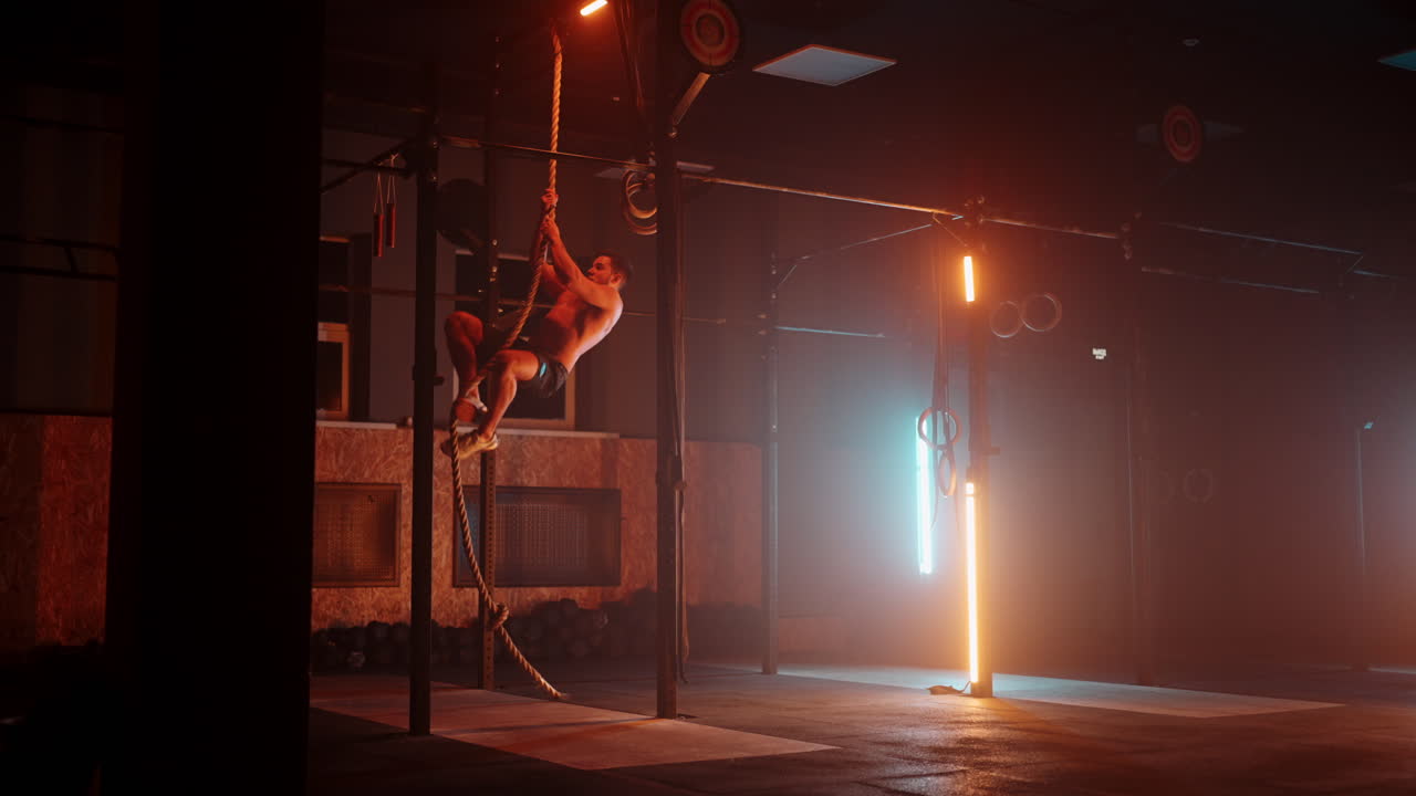 Uomo fitness che esegue un esercizio di arrampicata su corda in palestra al rallentatore. Luce al neon arancione e blu. Uomo che si allena in palestra arrampicandosi su una corda.