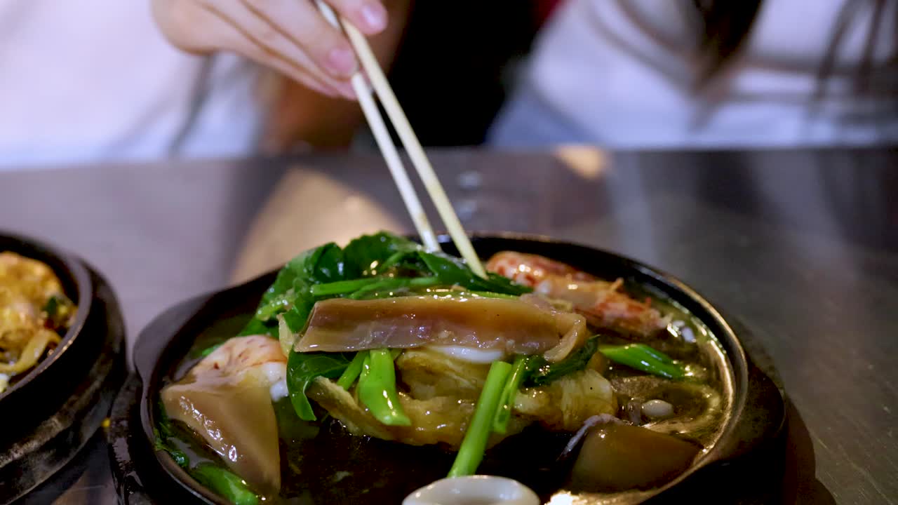 Woman uses chopsticks to pick up noodles and vegetables from steaming hot plate under bright lighting