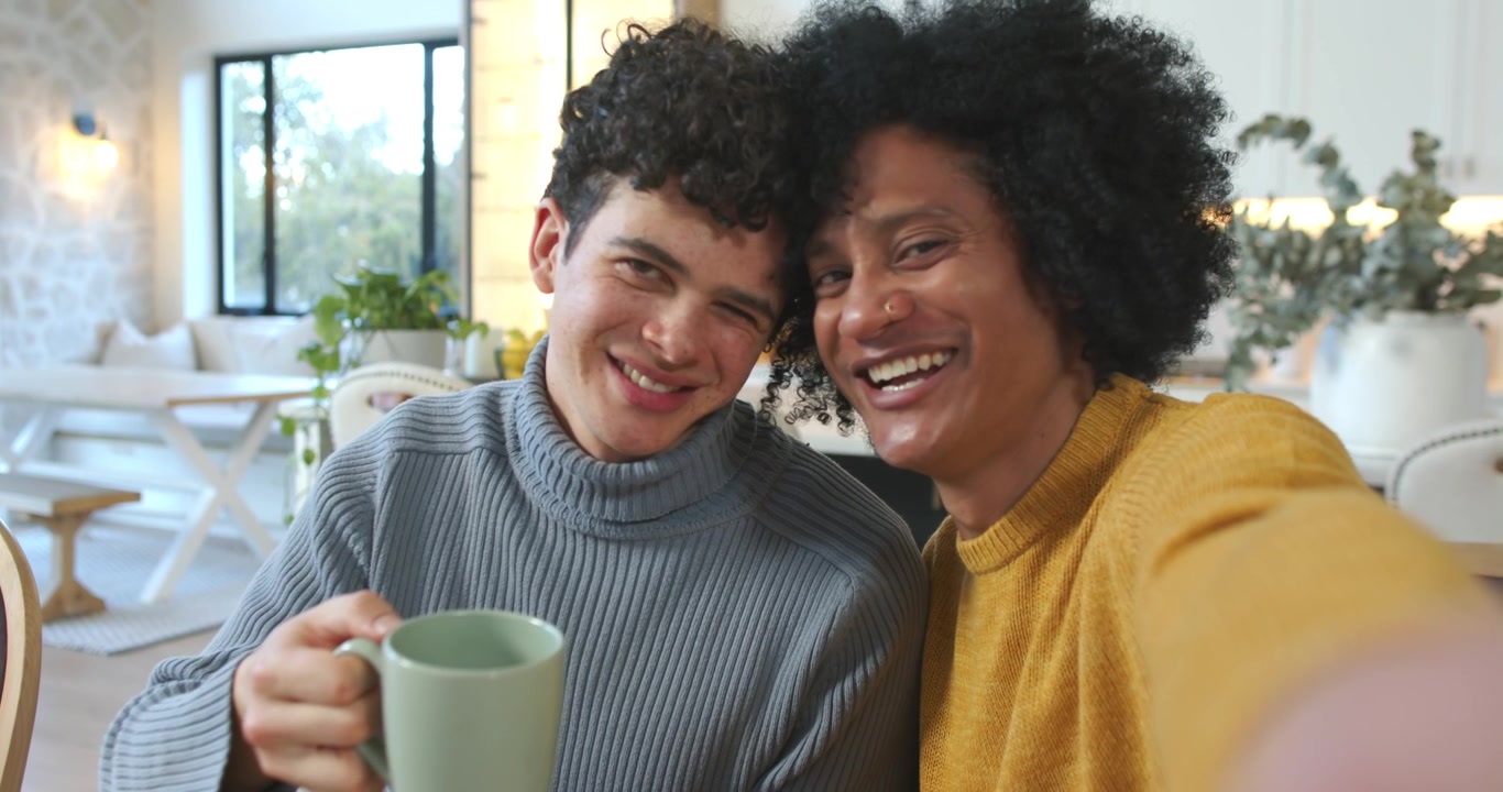 Two men holding smartphone taking selfie, clinking mugs sharing affection at dining table