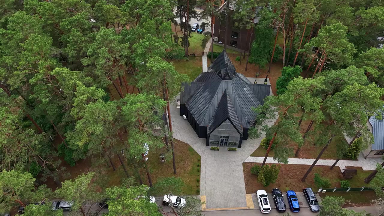 Betagende dronevideo af Kulautuva Kirke midt mellem høje fyrretræer og parkerede biler, der viser unik moderne arkitektur, rolige omgivelser og blødt dagslys fra luften