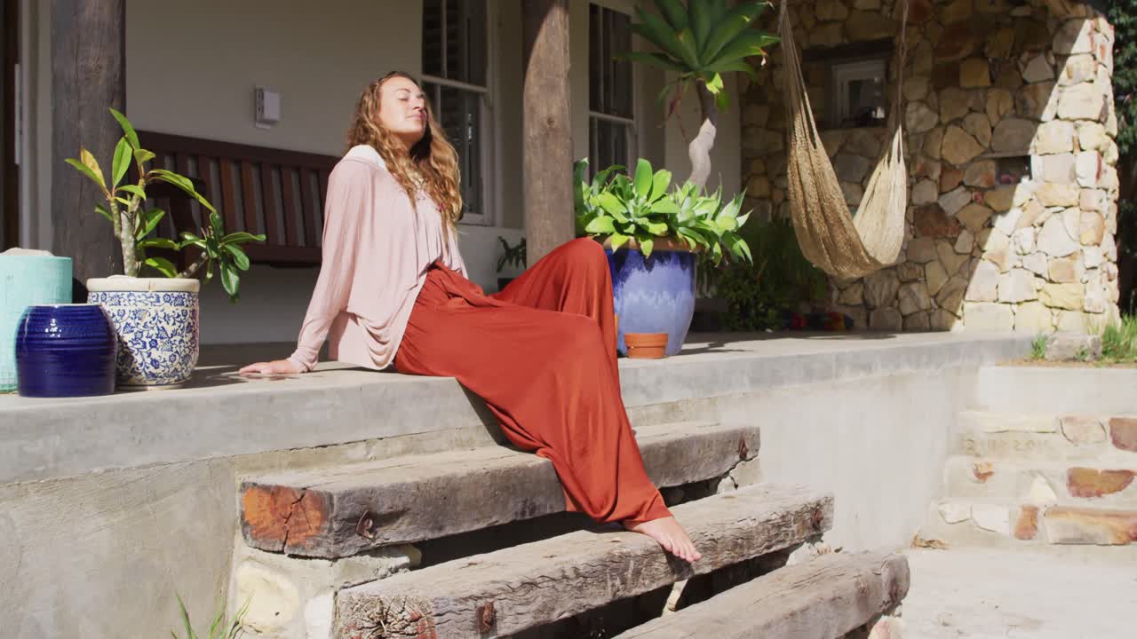 feliz mujer caucásica sentada en los escalones, relajándose con los ojos cerrados en la terraza de la cabaña soleada
