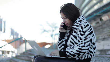 Concentrated-brunette-lady-talking-on-phone-and-looking-at-laptop