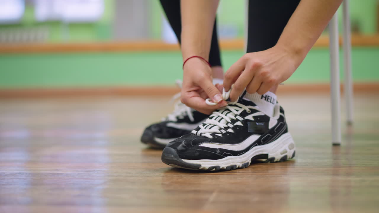 Vista delle gambe di una persona che indossa leggings neri, seduta su una sedia mentre allaccia le stringhe di scarpe da ginnastica di tela bianche e nere su un pavimento di legno; le mani tirano le stringhe con decisione, un braccialetto rosso è visibile al polso.