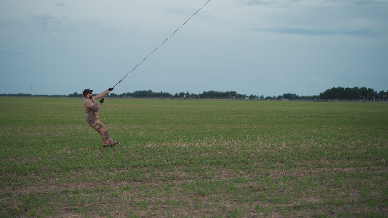man holding rope to anchor colorful hot air balloon against breeze on expansive green field under overcast sky capturing tension and preparation for flight in rural open
