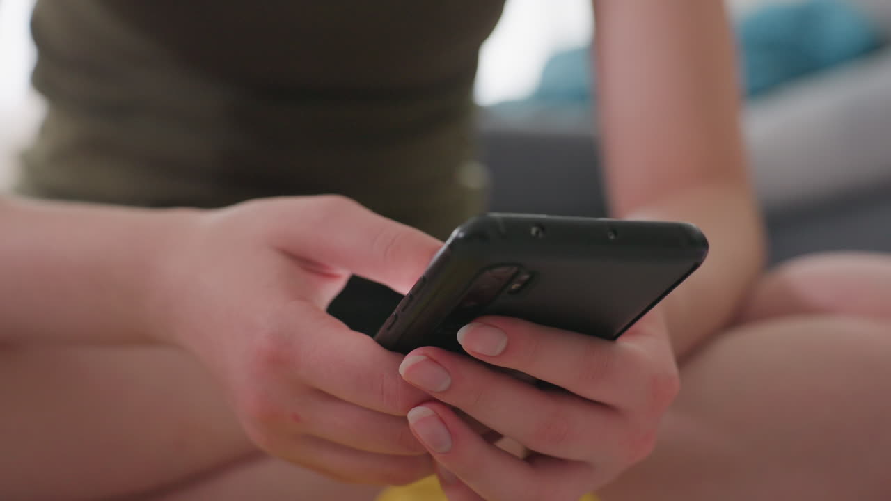 Close up partial view of fair skin person seated indoors using smartphone with both hands, fingers scrolling illuminated screen while relaxed posture conveys calm mood