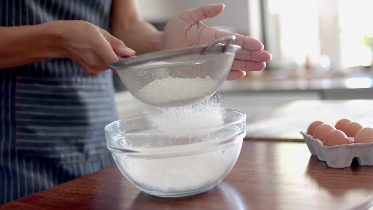 sección media de una mujer biracial en delantal tamizando harina, horneando en la cocina, cámara lenta