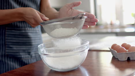 midsection of biracial woman in apron sieving flour, baking in kitchen, slow motion