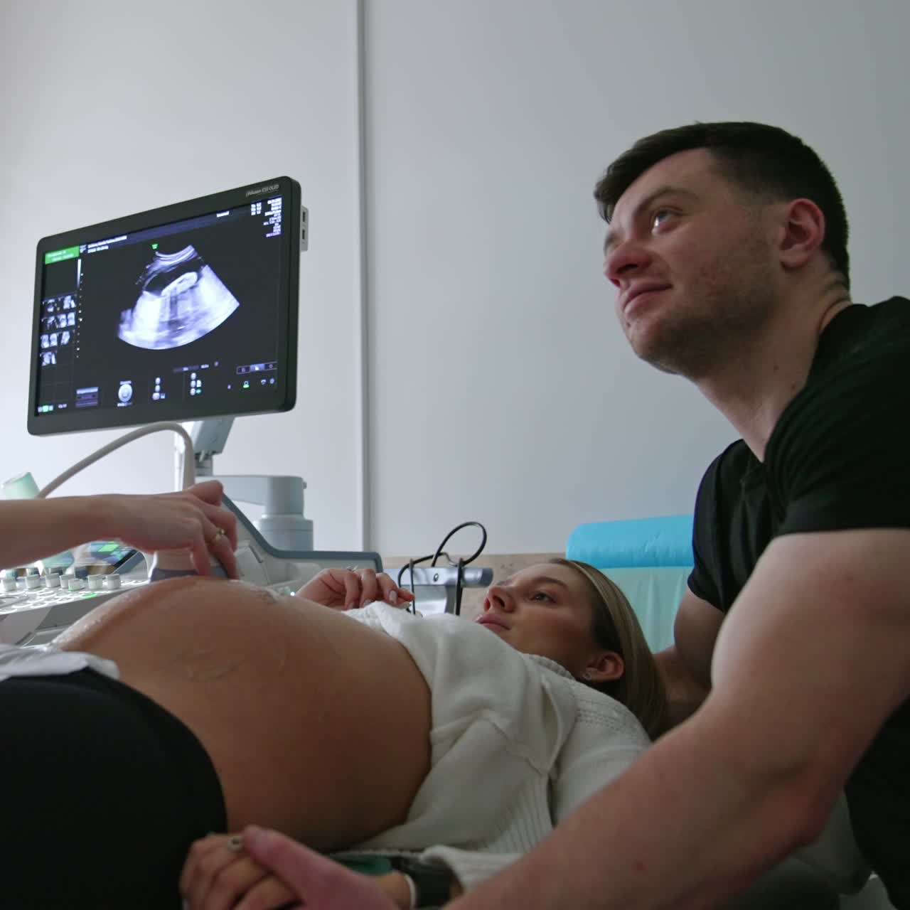 Pregnant wife and her husband are at the doctor's cabinet. Female doctor moving the sensor by the big belly of a woman