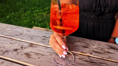 female hand holds red drink over wooden table outdoors, warm casual setting in natural light