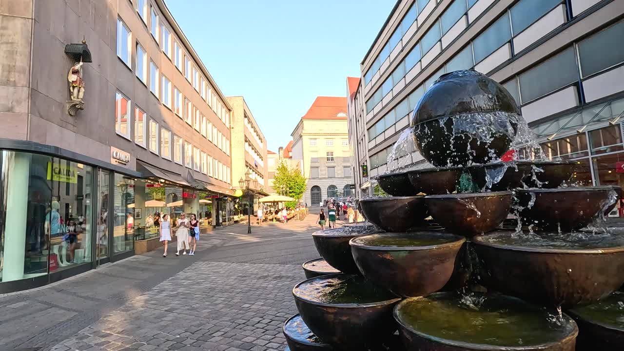 A multi-tiered stone fountain with a spherical top flows in a sunlit urban street, surrounded by modern buildings and pedestrians. Steady wide-angle camera