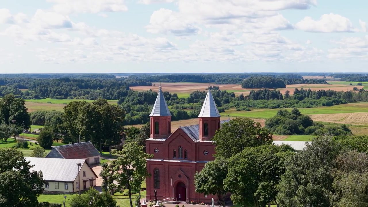 Flygfoto fångar Šatiai Johannes Döparens kyrkas röda tegeltorn mitt i frodig bygrönska, böljande litauiska fält och vidsträckta sommarhimlar i mjukt dagsljus. Ingen person i bild