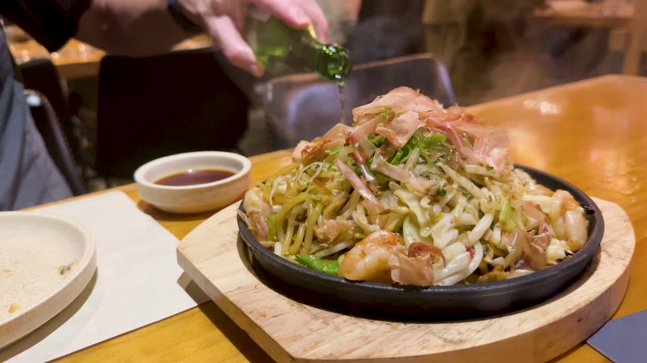 A hot plate of Japanese noodles garnished with fresh herbs, captured in a cozy restaurant setting with warm lighting