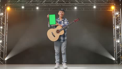 full body of asian teenager with guitar smiling and showing green screen tablet while standing on stage