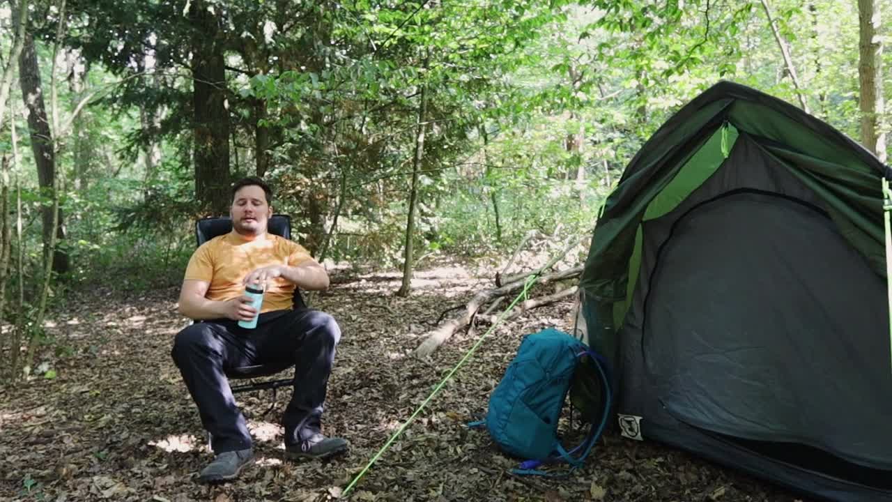 el hombre cansado y agotado se sienta en una silla frente a una carpa para descansar y beber agua