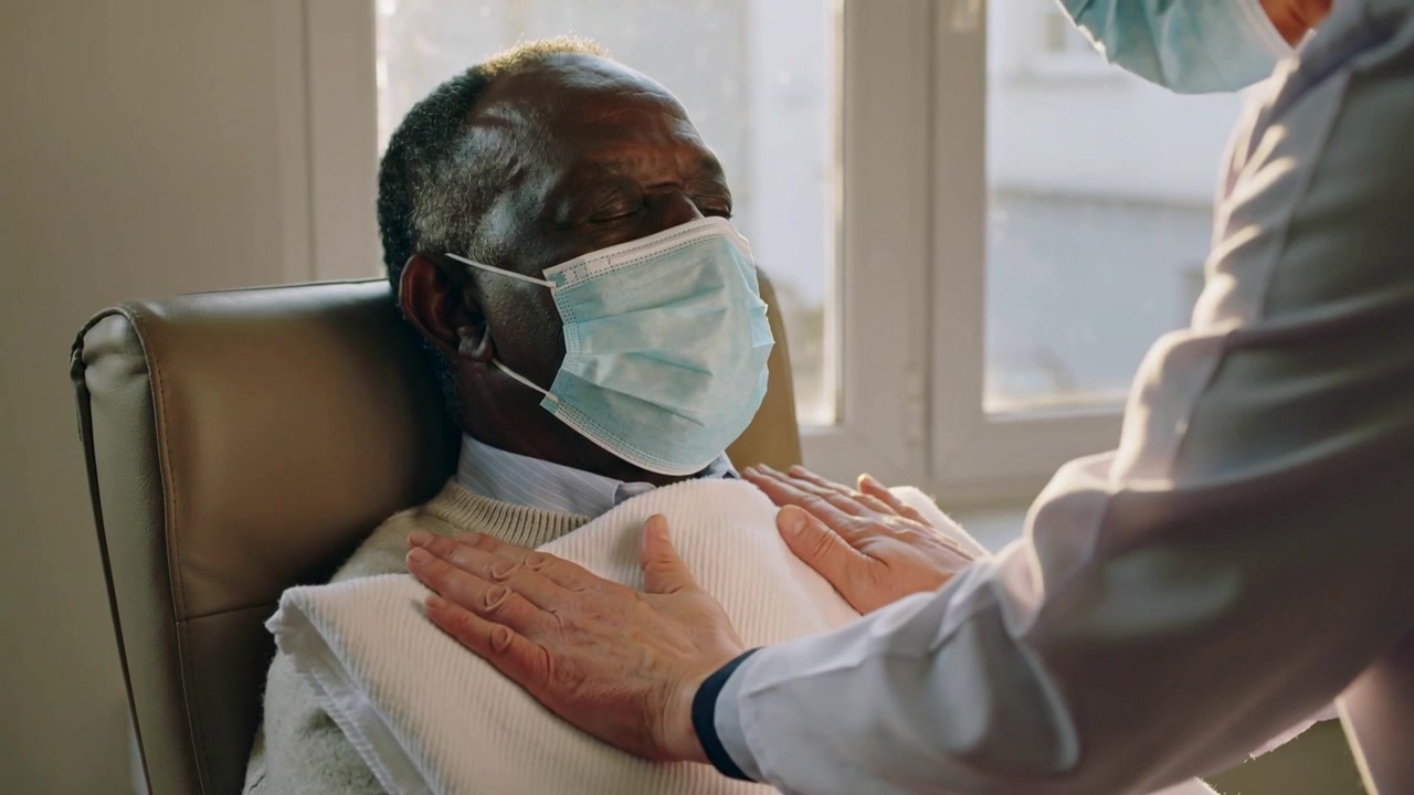 Perawat membentangkan handuk, menyelimuti lansia di kursi klinik sambil menepuk-nepuk untuk kehangatan, terlihat masker dan jas medis.