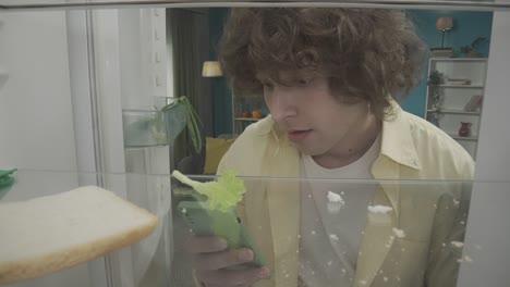 young man looking inside a messy fridge, talking on the phone