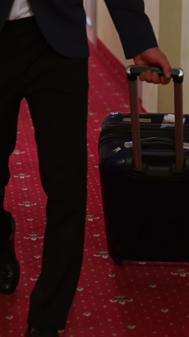 Young businessman is pulling suitcase. Travelling guy wearing elegant costume walking with his luggage in hotel hallway. Slow motion. Vertical video