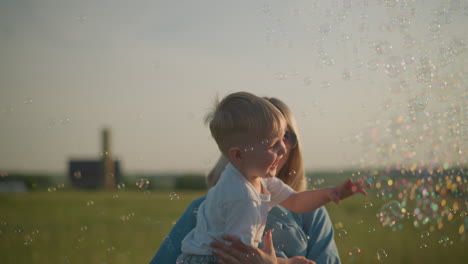 a joyful woman in a blue dress holds a little boy wearing a white shirt, as they delight in colorful bubbles floating around them in a grassy field, creating a magical and playful moment
