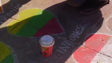 medium shot of a person drawing hearts with chalk on the the sidewalk with a cup of coffee sitting on the ground, shadow, handheld