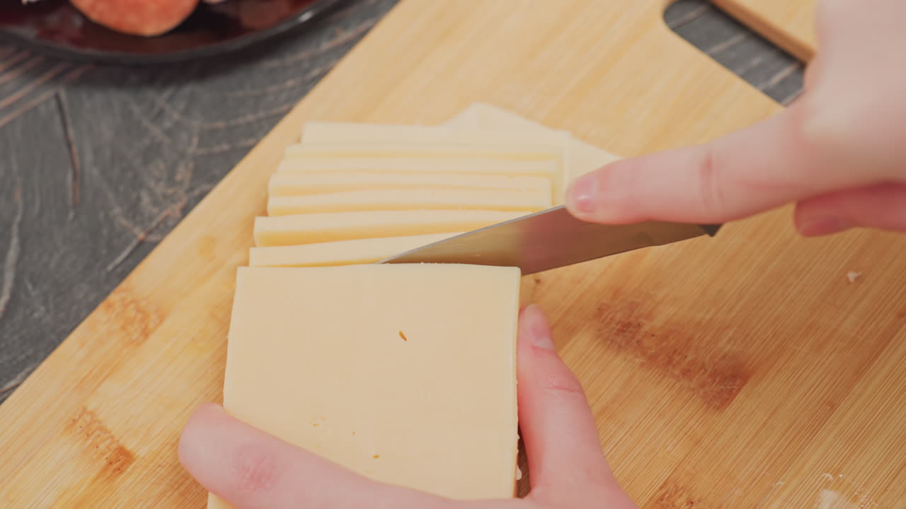 close up of person slicing block butter into even slices using sharp knife on wooden cutting board during food preparation in kitchen, showing detail of hands, blade, and butter texture in action