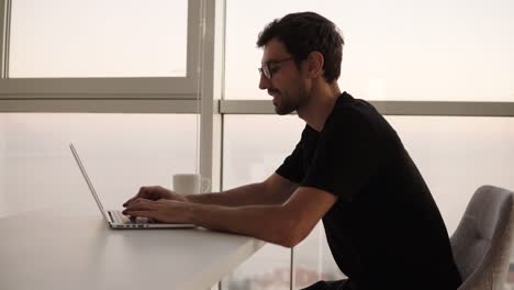 Side-view-of-business-man-using-laptop-computer-at-remote-workplace-in-slow-motion.-Freelancer-working-on-computer-at-home.-Focused-man-looking-on-screen-and-actively-typing-at-big-house-with-panoramic-windows.-Close-up