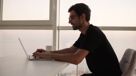 side view of business man using laptop computer at remote workplace in slow motion. freelancer working on computer at home. focused man looking on screen and actively typing at big house with panoramic windows. close up