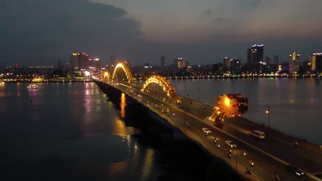 Drone-aerial-flying-towards-head-of-Dragon-Bridge-Cau-Rong,-traffic-and-city-skyline-at-night-in-Danang,-Vietnam