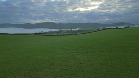 Flying-over-empty-road-with-rise-revealing-green-patchwork-fields,-mist-in-valley-and-distant-mountains-at-sunrise