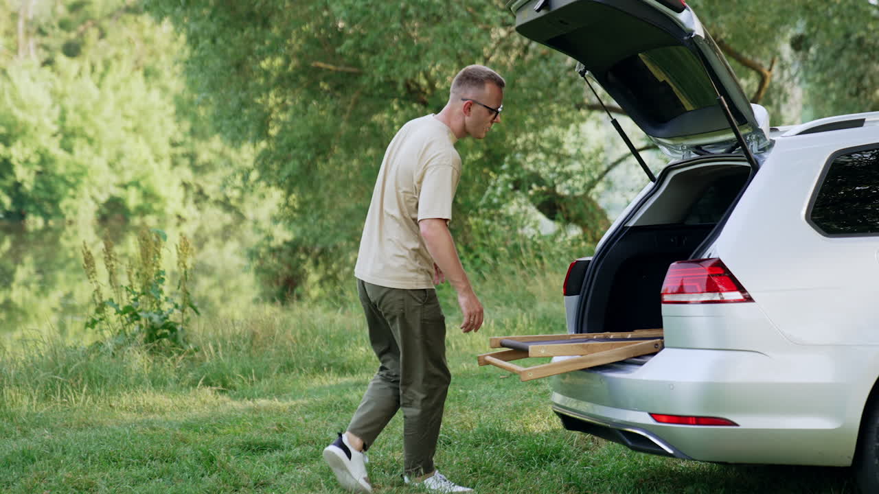 Man in beige t-shirt and khaki pants walks up to his car. Man takes folding chair out of the trunk. Nature at backdrop.