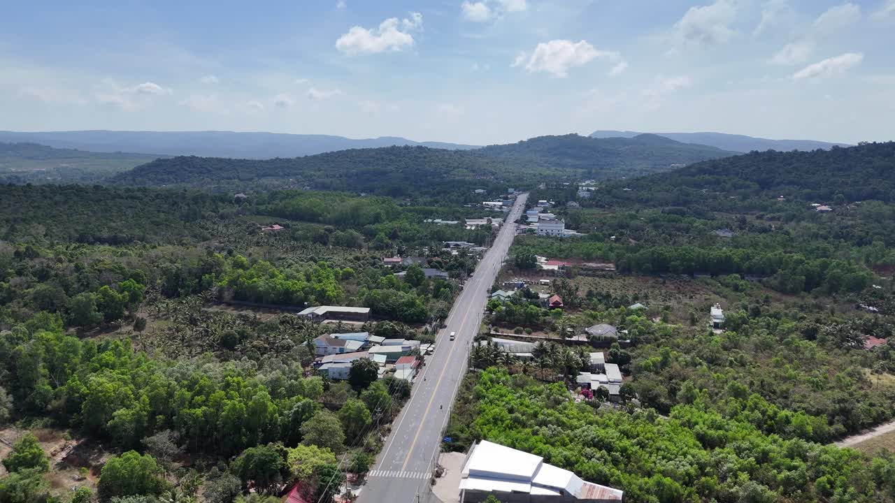 Aerial footage shows a main road greenery on either side and residents visible at intervals, creating a calm and peopled street. Passing clouds move overhead, and their shadows play across the road.