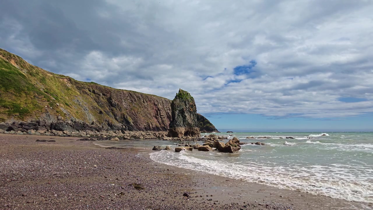 Irlands episke steder: blide bølger på stenstrand, klipper og havstak uden mennesker, vild natur i Waterford på en solrig sommerdag.