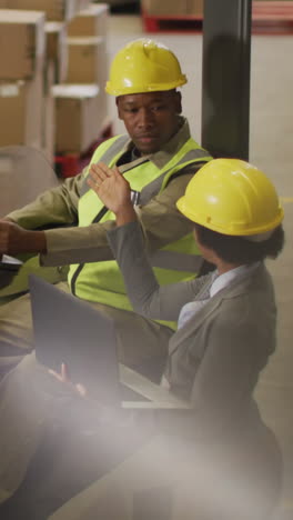 african american male and female workers wearing safety suits and using laptop in warehouse