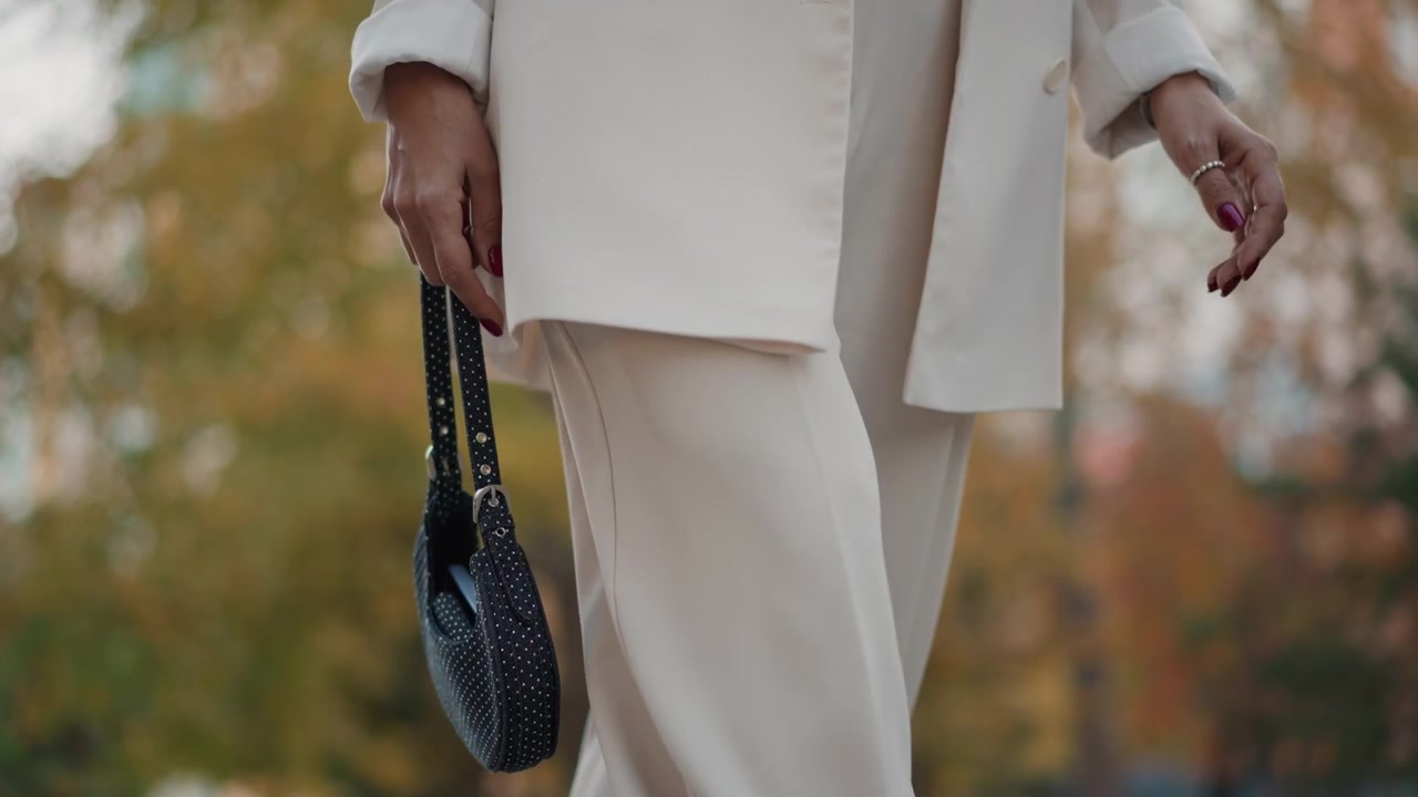 Vista dalla vita alla coscia di una donna che cammina all'aperto, tenendo una piccola borsa borchiata per il cinturino intrecciato mentre la borsa oscilla accanto a un blazer sartoriale e pantaloni cropped alla luce soffusa del giorno