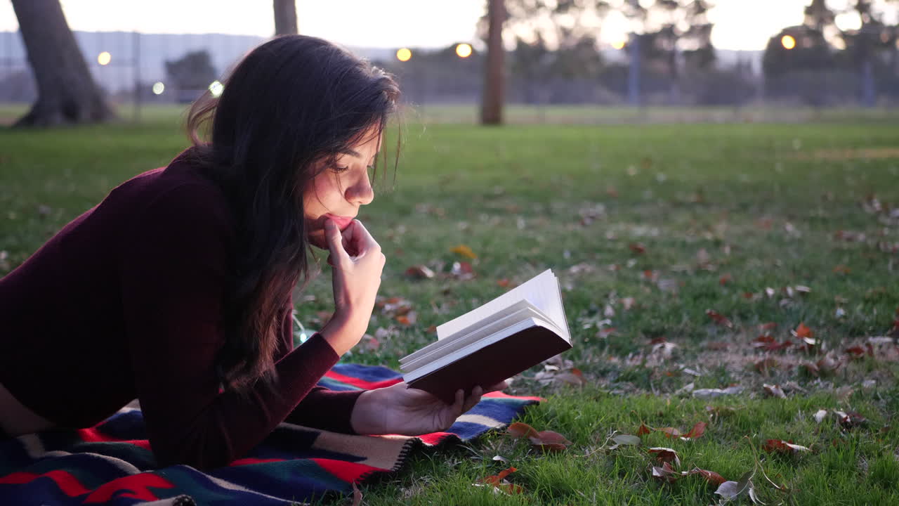 una estudiante hispana leyendo y estudiando un libro de texto escolar en un campus universitario al atardecer
