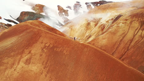 Hiker-Walking-On-The-Ridge-Of-Hveradalir-In-Kerlingarfjoll,-Iceland-With-Geothermal-Smoke-In-Background