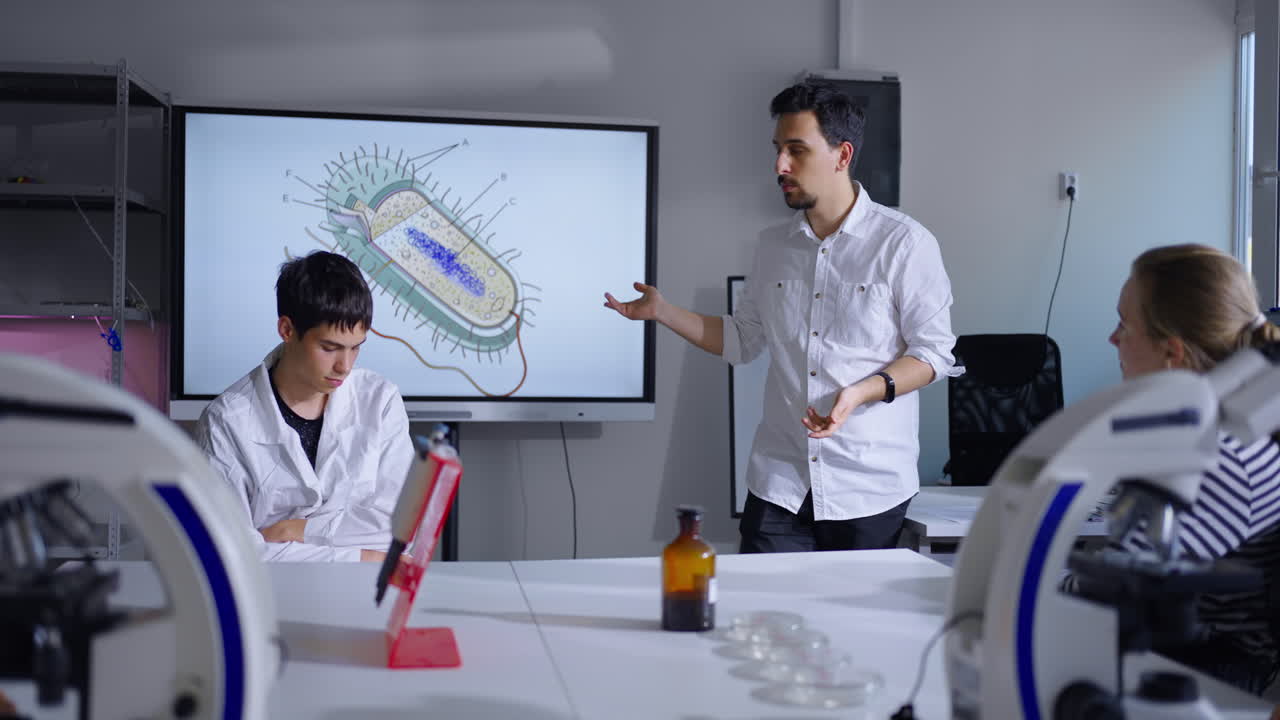 un profesor de biología da conferencias a los estudiantes en un aula con microscopios y un diagrama de una célula bacteriana en la pantalla