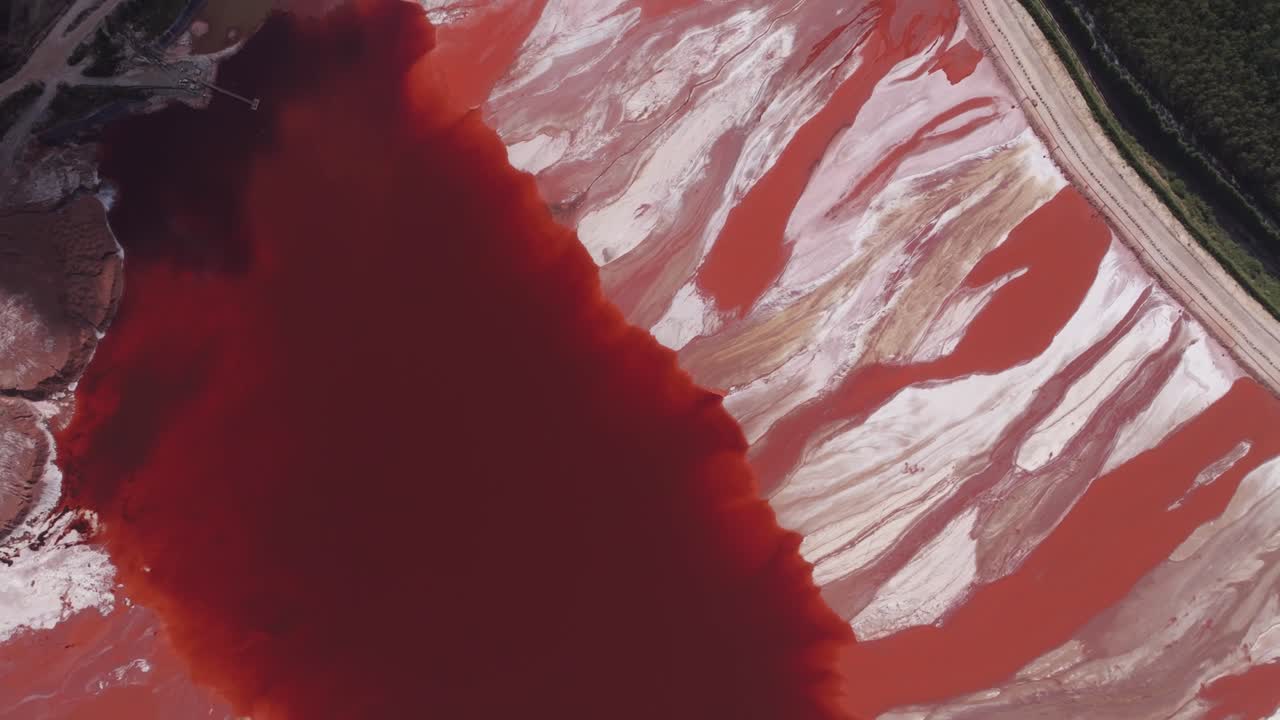 Aerial view of a massive red mud pond on the coast, an industrial waste byproduct from alumina production. The image highlights the enormous scale of the reservoir, with strong contrasts between the r